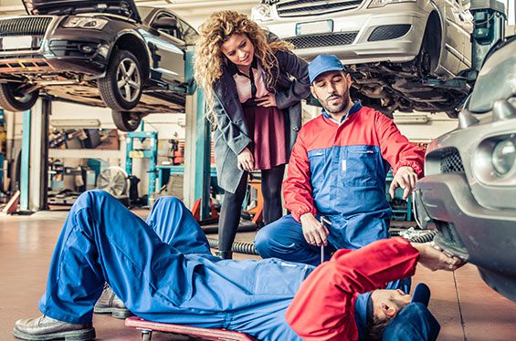 Two mechanics repair a car on a lift while the owner looks on